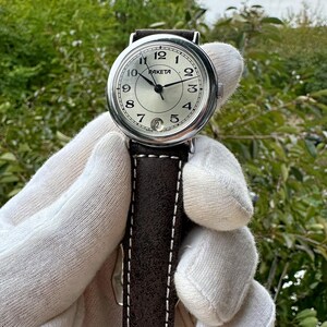 May include: A Raketa wristwatch with a silver dial, black hands, and a date window at the 6 o'clock position. The watch has a stainless steel case and a brown leather strap with white stitching. The watch is held against a backdrop of green foliage.