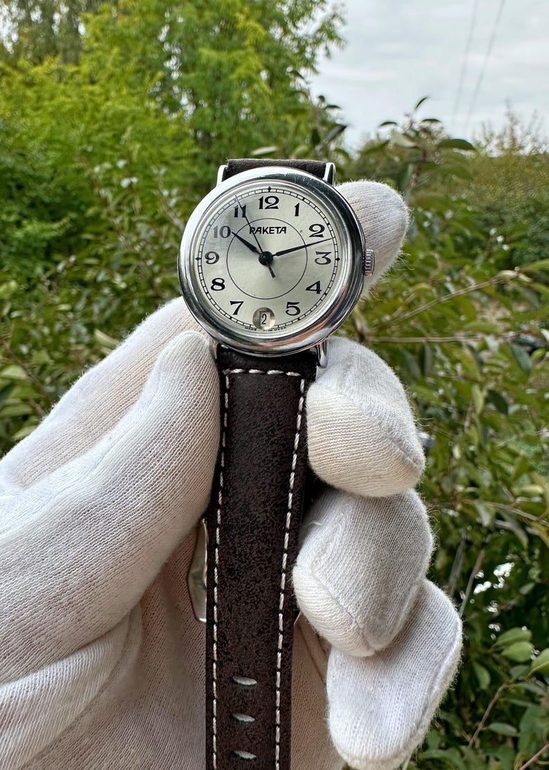 May include: A Raketa wristwatch with a silver dial, black hands, and a date window at the 6 o'clock position. The watch has a stainless steel case and a brown leather strap with white stitching. The watch is held against a backdrop of green foliage.