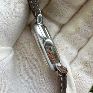 May include: Close-up of a silver wristwatch with a rectangular face and a brown leather strap. The watch has a small crown and a polished finish, reflecting the light. The strap is stitched and appears to be made of genuine leather.