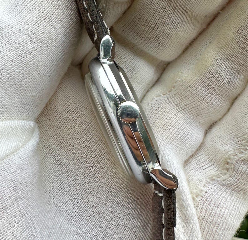 May include: Close-up of a silver wristwatch with a rectangular face and a brown leather strap. The watch has a small crown and a polished finish, reflecting the light. The strap is stitched and appears to be made of genuine leather.