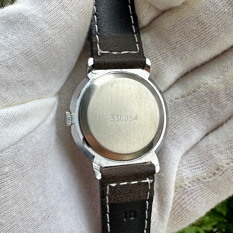 May include: Close-up of a vintage wristwatch, showcasing the back of the watch. The watch has a silver-colored case and a dark brown leather strap with white stitching. The watch face displays the number "336254".