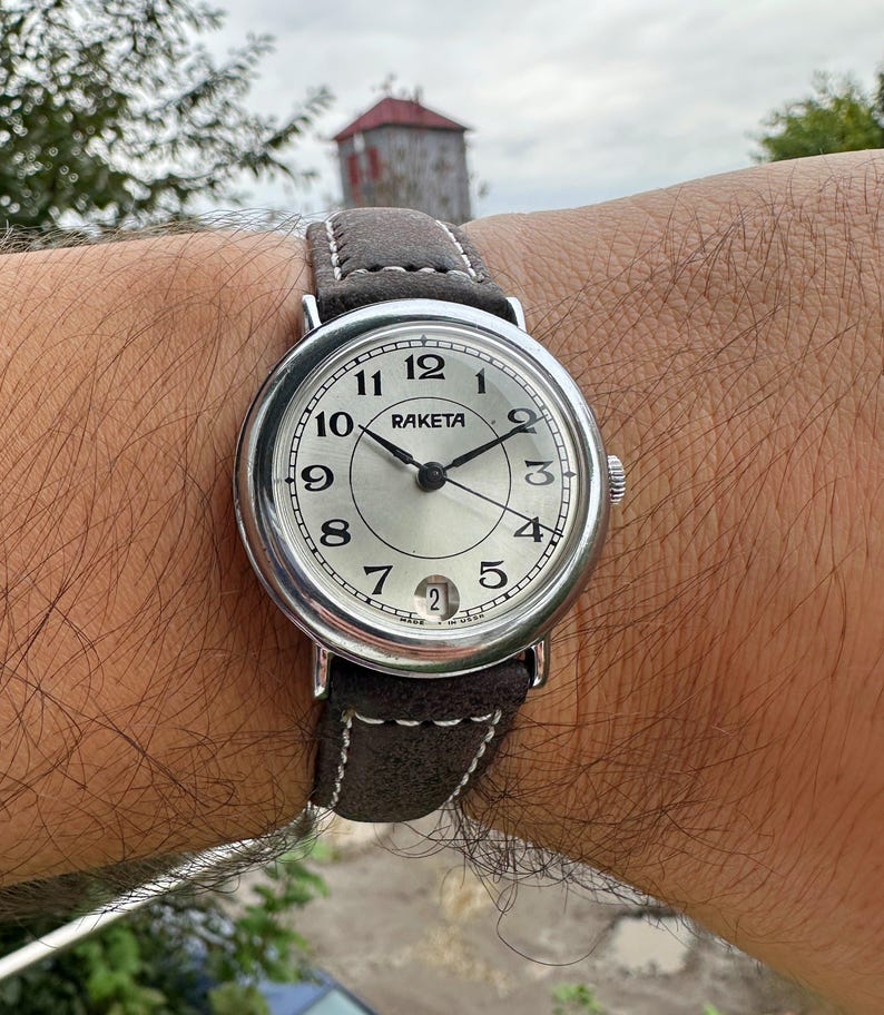 May include: A silver-toned Raketa wristwatch with a light-colored face and black hands. The watch has a brown leather strap with white stitching. The watch face displays the numbers 1 through 12, and the date. The watch is on a wrist.