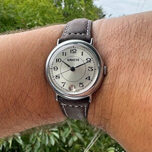 May include: A silver-toned Raketa wristwatch with a gray suede band. The watch face is silver with black numerals and hands. The watch is secured on a wrist.