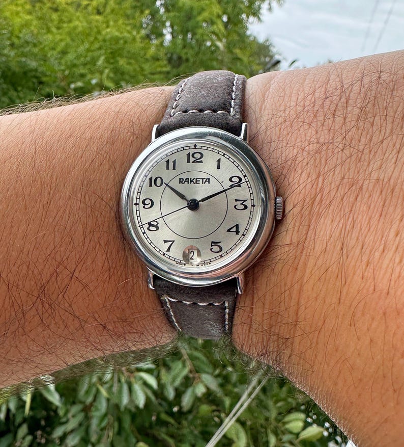 May include: A silver-toned Raketa wristwatch with a gray suede band. The watch face is silver with black numerals and hands. The watch is secured on a wrist.