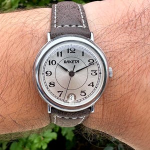 May include: A silver-toned Raketa wristwatch with a light-colored face and black numerals. The watch has a brown suede band and a date window at the 6 o'clock position. The watch hands are black.