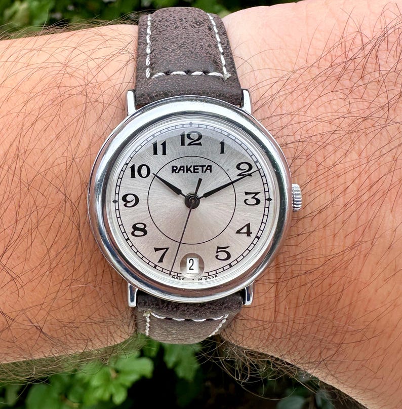 May include: A silver-toned Raketa wristwatch with a light-colored face and black numerals. The watch has a brown suede band and a date window at the 6 o'clock position. The watch hands are black.