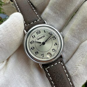 May include: A silver-toned Raketa wristwatch with a light-colored face, black hands, and a date window. The watch has a brown leather strap with white stitching. The watch face displays the numbers 1 through 12.