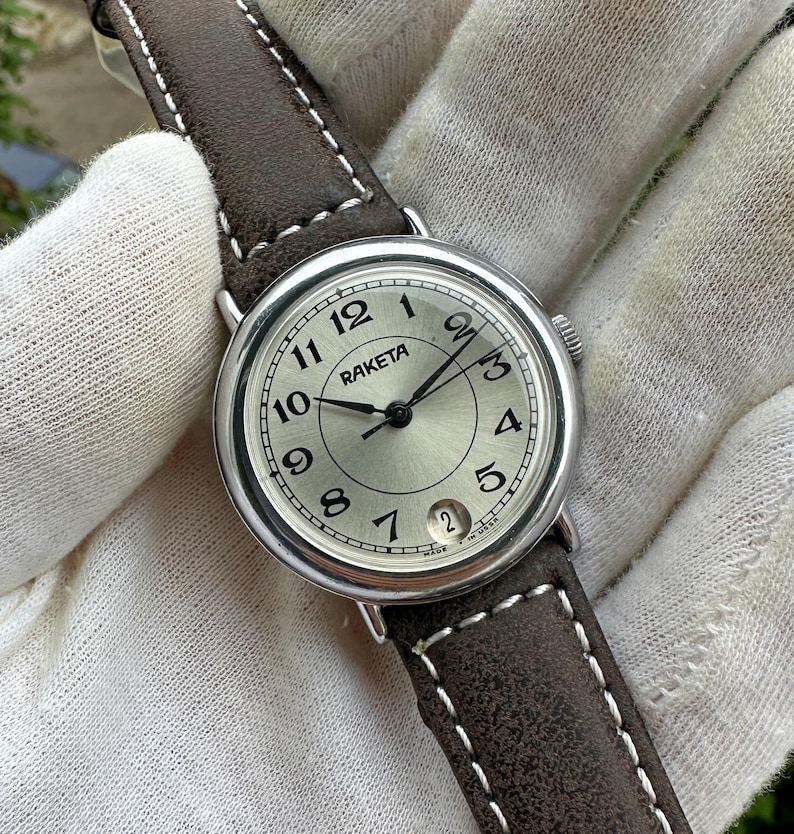 May include: A silver-toned Raketa wristwatch with a light-colored face, black hands, and a date window. The watch has a brown leather strap with white stitching. The watch face displays the numbers 1 through 12.