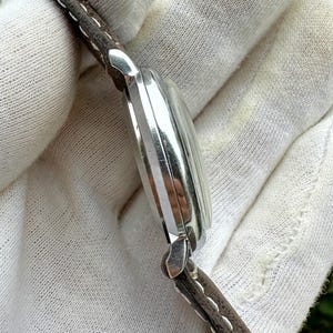 May include: A silver-toned wristwatch with a brown leather strap. The watch face is not visible, but the side profile shows a curved design with vertical lines. The strap is stitched with white thread.