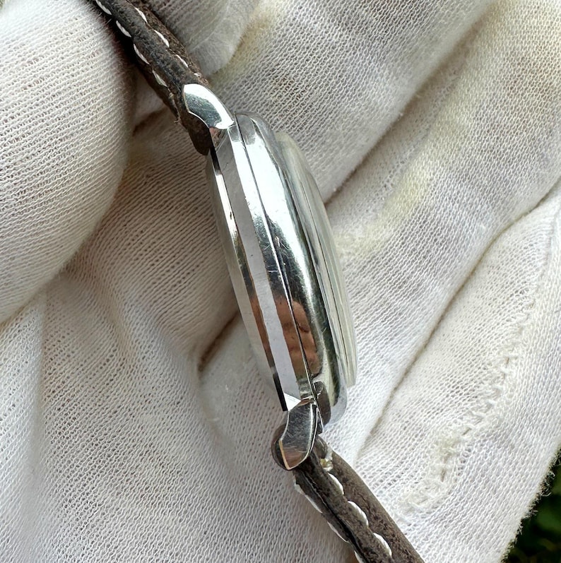 May include: A silver-toned wristwatch with a brown leather strap. The watch face is not visible, but the side profile shows a curved design with vertical lines. The strap is stitched with white thread.