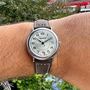 May include: A silver Raketa wristwatch with a light gray face, black hands, and a brown suede band. The watch face displays the numbers 1 through 12, with the brand name "Raketa" above the center. The band is approximately 20 mm wide.