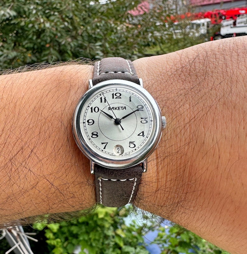 May include: A silver Raketa wristwatch with a light gray face, black hands, and a brown suede band. The watch face displays the numbers 1 through 12, with the brand name "Raketa" above the center. The band is approximately 20 mm wide.
