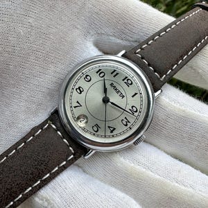 May include: A silver-toned Raketa wristwatch with a light-colored face, black hands, and black Arabic numerals. The watch has a brown leather band with white stitching. The date is visible at the 6 o'clock position.