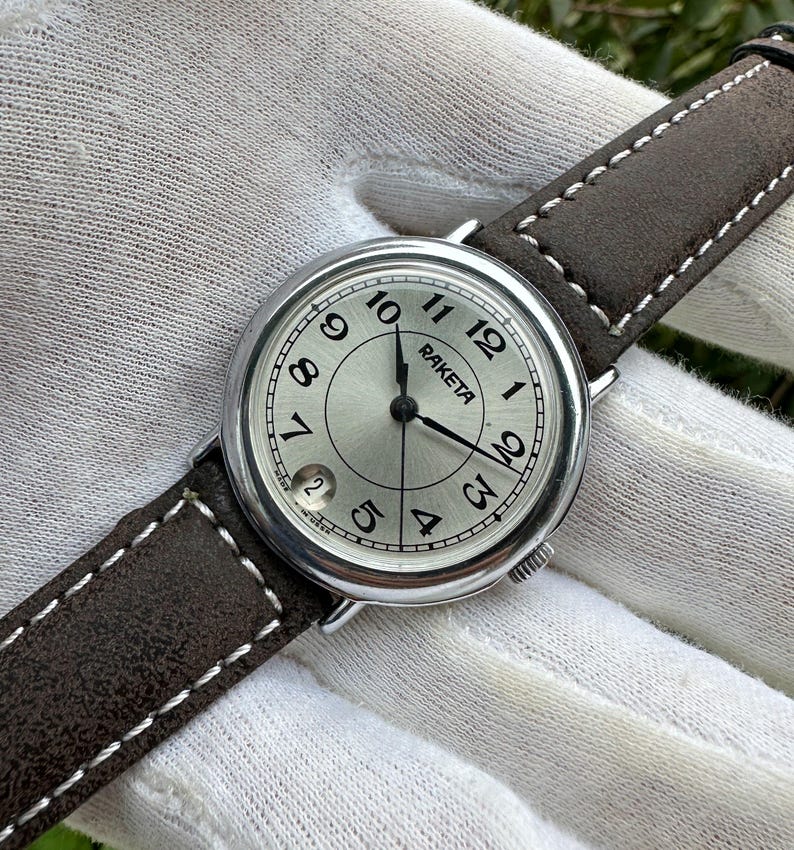 May include: A silver-toned Raketa wristwatch with a light-colored face, black hands, and black Arabic numerals. The watch has a brown leather band with white stitching. The date is visible at the 6 o'clock position.