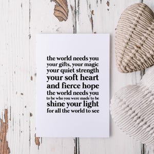 Op de afbeelding: Een witte kaart met zwarte tekst met de tekst "de wereld heeft je nodig...laat je licht schijnen voor de hele wereld". Twee hartvormige decoratieve objecten liggen naast de kaart. De achtergrond is een verweerd wit houten oppervlak.