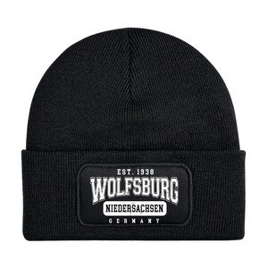 Può includere: Berretto nero a maglia con una toppa rettangolare con la scritta "WOLFSBURG" sopra "NIEDERSACHSEN GERMANY" e "EST. 1938" in bianco. Il berretto è in stile classico, con risvolto, adatto al freddo.
