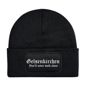 Könnte beinhalten: Schwarze Strickmütze mit einem rechteckigen Aufnäher. Der Aufnäher zeigt das Wort "Gelsenkirchen" in gotischer Schrift und den Satz "You'll never walk alone". Ein klassisches Winteraccessoire.