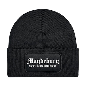 Könnte beinhalten: Schwarze Beanie-Mütze mit rechteckigem Aufnäher. Der Aufnäher zeigt das Wort "Magdeburg" in gotischer Schrift über dem Satz "You'll never walk alone". Die Mütze ist aus Strickmaterial.