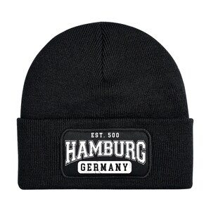 Op de afbeelding: Zwarte beanie-muts met een rechthoekige patch met de tekst "HAMBURG GERMANY" in het wit, met "EST. 500" erboven. De muts is gemaakt van gebreid materiaal, geschikt voor koud weer.