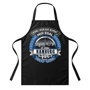 Könnte beinhalten: Schwarze Schürze mit einem grafischen Design mit dem Text "GEQUALT, AUSGELACHT, BESPUCKT. MIR EGAL" und "HAMBURG Fan". Das Design beinhaltet einen blauen Lorbeerkranz und eine Stadtlandschaft.