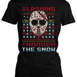 Könnte beinhalten: Schwarzes T-Shirt mit einem pixeligen Design, das eine weiße Hockeymaske mit roten Akzenten und die Worte "SLASHING THROUGH THE SNOW" in Rot und Blau zeigt. Das Design hat einen festlichen, hässlichen Sweater-Stil.