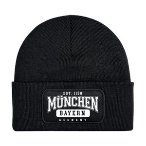 Può includere: Berretto nero con una toppa rettangolare con la scritta bianca "MUNCHEN", con "BAYERN" sotto e "GERMANY" in basso. Il testo "EST. 1158" è sopra. Il berretto è realizzato in tessuto a maglia.