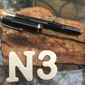 May include: A black fountain pen with a silver trim and a black cap. The pen is lying on a brown rock with the letters N3 written on it.