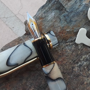 May include: A black and white marbled fountain pen with a gold trim and a silver nib. The pen is lying on a rock surface.