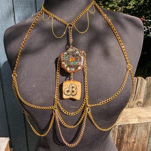 May include: A gold-colored body chain with a pendant featuring a colorful, abstract design. The chain drapes across the shoulders and chest, with a second pendant below. The lower pendant has a carved design. The jewelry is displayed on a black mannequin.