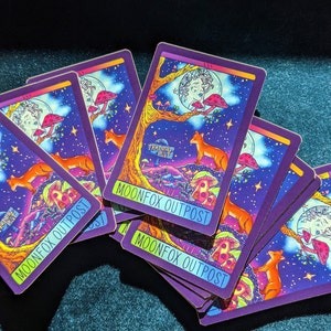May include: A stack of playing cards with a purple background and a colorful illustration of a fox, mushrooms, and a moon. The cards have the text "Moonfox Outpost" printed on them.