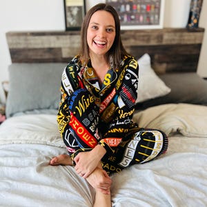 Puede incluir: Una persona vistiendo un pijama negro y blanco con un estampado colorido de palabras y logotipos. El pijama tiene una camisa abotonada y pantalones largos. La persona está sentada en una cama blanca con una cabecera de madera.