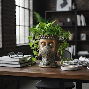 Könnte beinhalten: Ein bronzefarbener Buddha-Kopf-Pflanztopf mit dekorativer Gesichtsmaske, gefüllt mit grünen Blättern. Der Pflanztopf steht auf einem Holztisch, daneben eine Brille und Bücher. Der Hintergrund umfasst ein Fenster und ein Bücherregal.