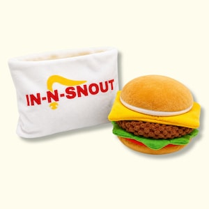 May include: A white plush pouch with the red text IN-N-SNOUT and a yellow graphic, paired with a plush hamburger toy featuring a tan bun, a yellow cheese slice, a brown textured patty, and a green lettuce layer, all made of soft fabric.
