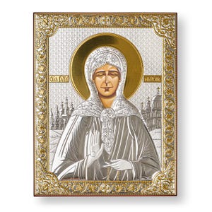 May include: A religious icon featuring a woman with a gold halo, wearing a white head covering and silver robes. The icon is set within a gold and silver decorative frame, with a brown wooden border. The background includes a church and the name "MATPOHA".
