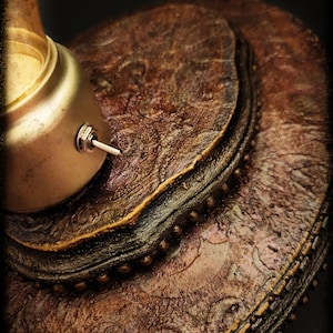 May include: A close-up of a lamp base with a textured, layered, brown and gold finish. The base has a round shape and a small switch on the side.