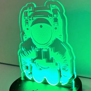 May include: A green astronaut-shaped night light with a black base. The astronaut is wearing a helmet and has a detailed design.