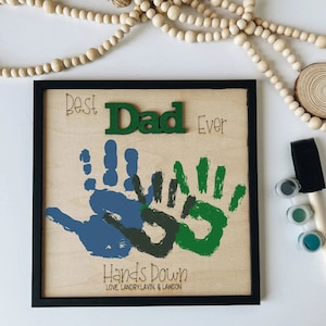 May include: A framed wooden sign with the words "Best Dad Ever" and "Hands Down" in black lettering. The sign also features two handprints in blue and green paint. The sign is surrounded by a black frame.