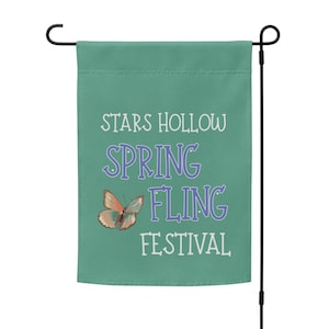 Puede incluir: Una bandera de jardín turquesa con las palabras "STARS HOLLOW SPRING FLING FESTIVAL" en letras blancas y moradas. También presenta un gráfico de mariposa. La bandera está sujeta a una estaca de jardín de metal negro.