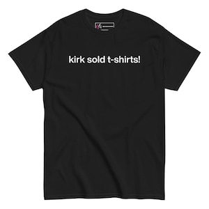 May include: A black short-sleeved t-shirt featuring the white text 'kirk sold t-shirts!' printed across the chest. The garment includes a small rectangular logo tag on the inside of the neckline with the letters 'GG' in pink and white.