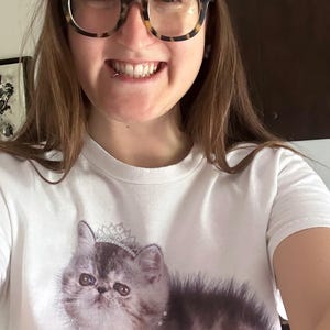 May include: A white t-shirt featuring a graphic of a fluffy gray cat wearing a crown and necklace, with the word "PRINCESS" in pink. The person is wearing a pink baseball cap with "TWICKHAM 2005" and tortoise shell glasses.