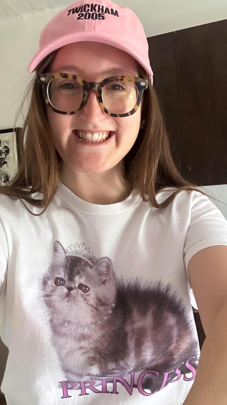 May include: A white t-shirt featuring a graphic of a fluffy gray cat wearing a crown and necklace, with the word "PRINCESS" in pink. The person is wearing a pink baseball cap with "TWICKHAM 2005" and tortoise shell glasses.