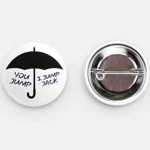Pode incluir: Dois botões redondos brancos. Um apresenta um guarda-chuva preto com o texto azul "You Jump, I Jump Jack". O outro botão mostra a parte de trás com um alfinete de metal.