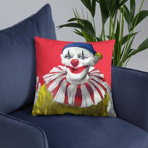 May include: A red throw pillow with a colorful clown face illustration. The clown is wearing a blue and yellow hat, a white and red striped collar, and a yellow shirt.