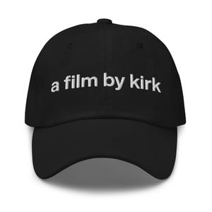 May include: Black baseball cap with a curved peak and adjustable strap. The cap has white embroidered text reading "a film by kirk" across the front. The cap is made of a soft material.