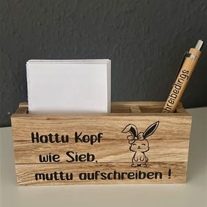 Könnte beinhalten: Ein rechteckiger Schreibtisch-Organizer aus Holz mit einem Stapel weißer quadratischer Notizzettel und einem Bambuskugelschreiber. Die Vorderseite zeigt eine schwarze Grafik eines Hasen mit Schleife und den Text: Hattu Kopf wie Sieb, muttu aufschreiben!