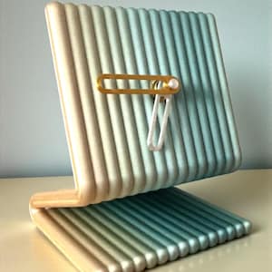 Könnte beinhalten: Ein moderner, abstrakter Schreibtisch-Organizer in einem Farbverlauf von Beige bis Türkis. Das Design weist eine gerippte Textur und einen goldfarbenen Akzent mit einem weißen Clip auf. Der Organizer ist zur Aufnahme von Gegenständen konzipiert.