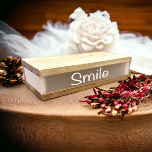 Könnte beinhalten: Eine Holzkiste mit weißem Hintergrund und dem aufgedruckten Wort "Smile". Die Kiste steht auf einer braunen Oberfläche mit einem weißen Stoff und getrockneten Blumen.