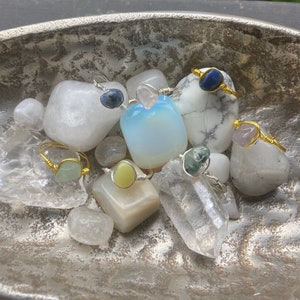 May include: A collection of rings featuring various gemstones, including blue, green, yellow, and pink, set in gold wire and displayed on a silver tray with white stones.