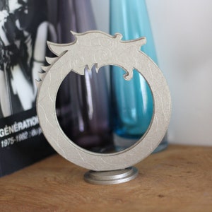 Op de afbeelding: Een zilverkleurige decoratieve standaard in de vorm van een drakenkop met een ronde opening. De standaard staat op een houten oppervlak.