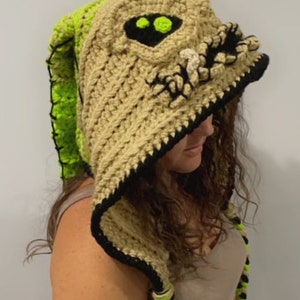 May include: A crocheted hooded hat with a beige and green color scheme. The hood features a detailed crocheted face with black eyes and a toothy grin. The hat has long, crocheted ties.